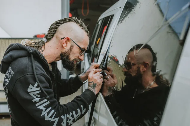 A man with dreadlocks applies tint to a vehicle window, focused and precise. NYC commercial vehicle wraps: FMCSA marking, TLC decals and tint records